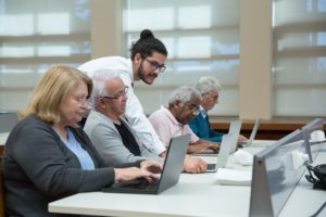 Man helping 4 elderly people at a table using laptops