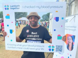 Jason smiling and wearing a hat at the Lambeth Country Show. He holds a sign which reads 'I've checked my blood pressure.'