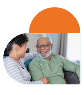 An elderly man sits on a sofa with a younger woman, his carer, both are smiling