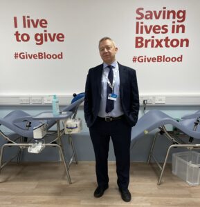 Andrew Eyres from NHS SOuth EAst London and Lambeth Council stands in fron of sign that sauys save lives give blood at launch of new Brixton Blood Donor Centre