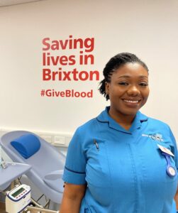 smiling Black woman in NHS unifrom stands in front of a sign that reads Saving lives in Brixton at Brixton Blood donor centre