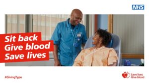 A blood donor sits in chair ready to give blood. She smiles at member of staff who is helping her get set up. Test says Sit back and give blood