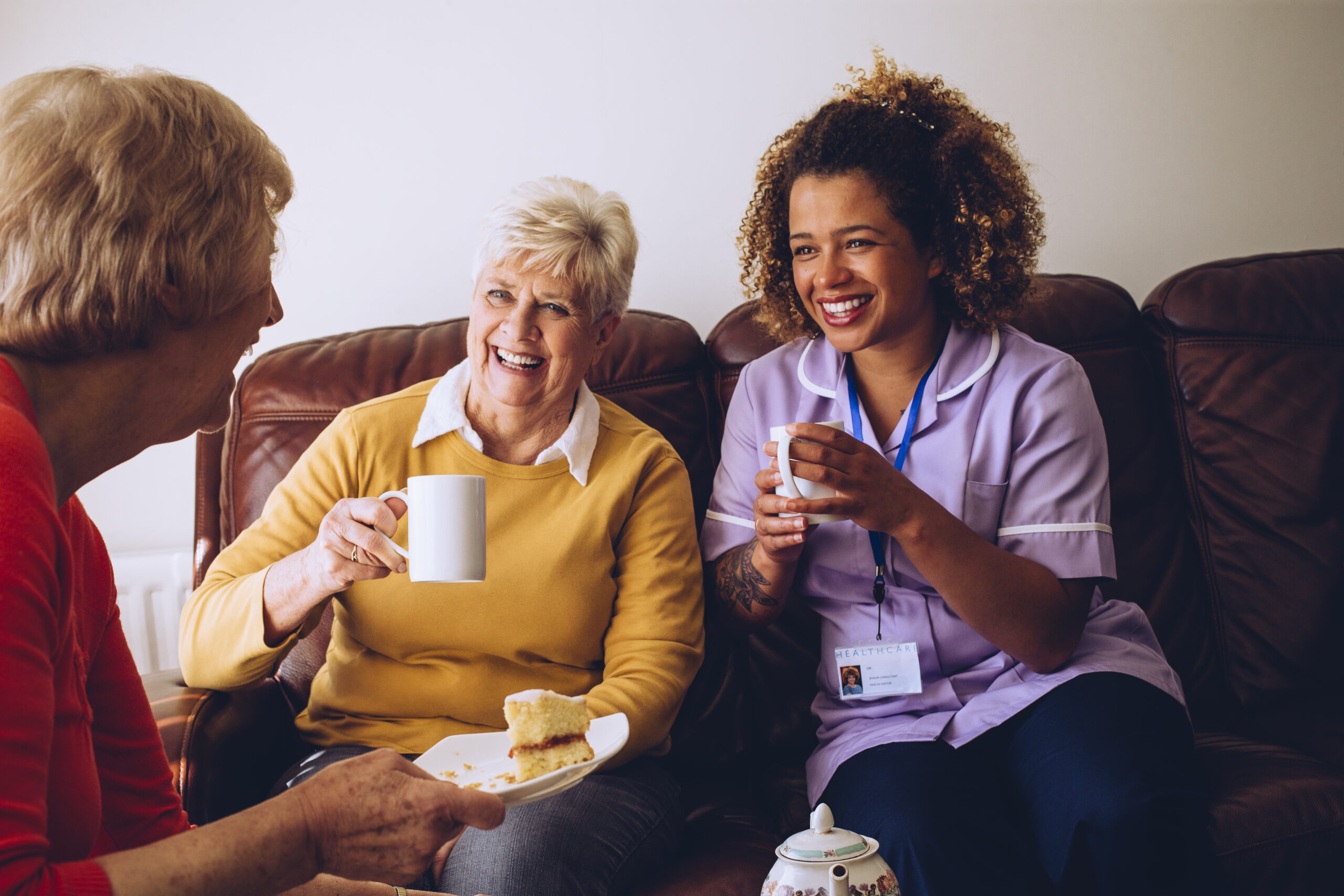 Elderly carer sitting with two of her patients in the care home, enjoying some cake and tea.